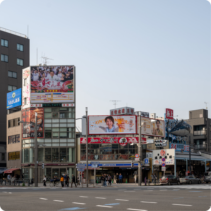 Un paysage urbain japonais avec des publicités colorées pour de la nourriture et des boissons, un magasin Lawson au centre et une grande sculpture de poisson sur le bâtiment à droite, sous un ciel clair en journée.