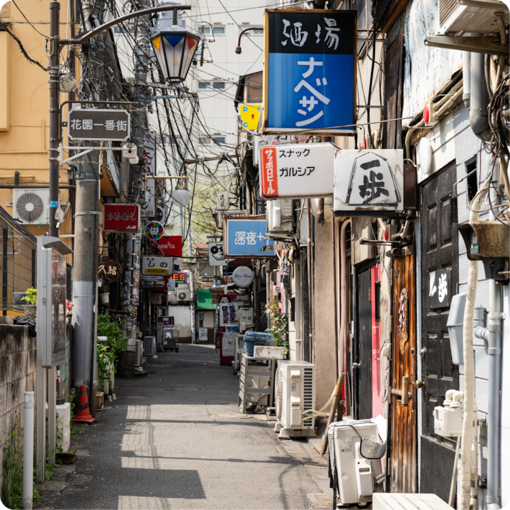 Une ruelle japonaise animée avec des enseignes de magasins colorées, des câbles électriques apparents et de petites devantures de magasins rapprochées, reflétant une atmosphère locale vivante et authentique.