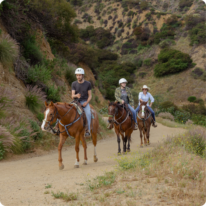 Trois personnes à cheval sur un sentier de terre entouré de verdure et de collines, pendant la journée.