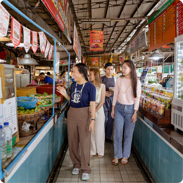 Quatre personnes se promènent dans un marché animé avec des étals de fruits, légumes et jus de fruits de chaque côté, éclairés par des lampes suspendues.