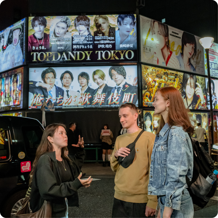 Un groupe de trois personnes discutent dans une rue animée de Tokyo la nuit, entourées de publicités lumineuses colorées pour la vie nocturne.