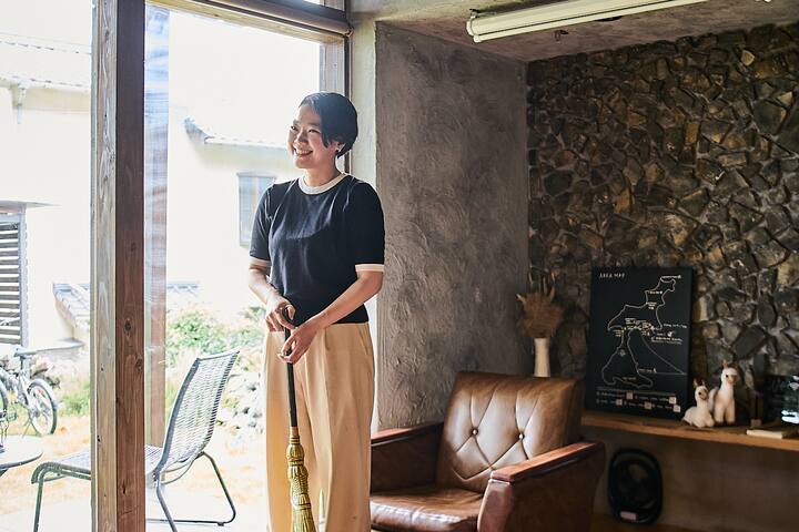 Person standing indoors near a large window, holding a broom, with a view of a garden outside and a cozy seating area featuring a leather chair and a stone wall with a map of Japan displayed.