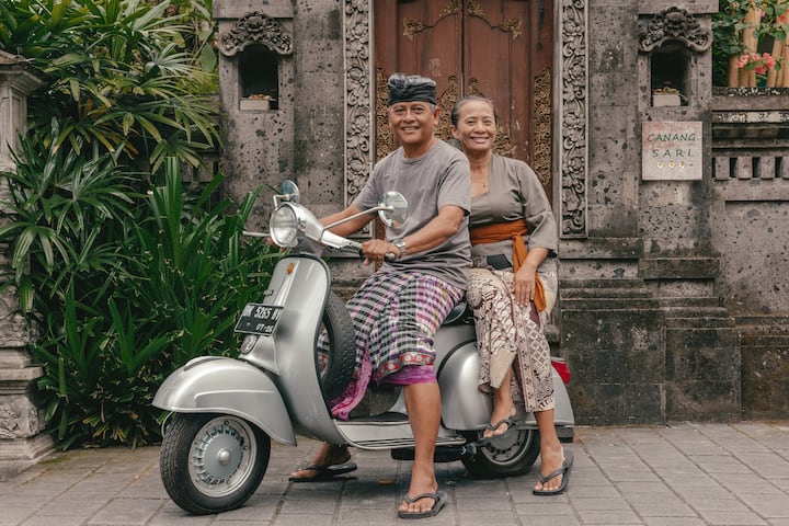 A man and a woman seated on a silver scooter in front of a traditional Balinese stone doorway surrounded by greenery.