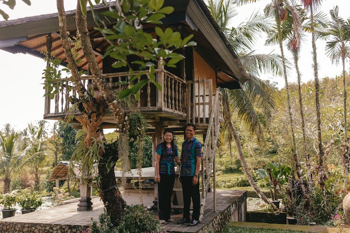 A traditional wooden villa with a balcony surrounded by lush greenery, palm trees, and tropical vegetation in Bali, Indonesia.