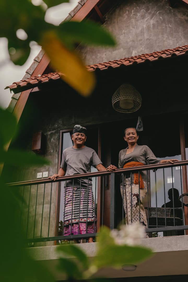 Two people dressed in traditional Balinese attire standing on a balcony of a rustic house with a red-tiled roof, surrounded by greenery.