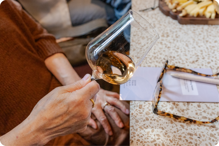 Une personne tient un verre de vin blanc pendant une dégustation, à côté d'une table avec un plateau de fromages, une fiche d'informations et des lunettes.