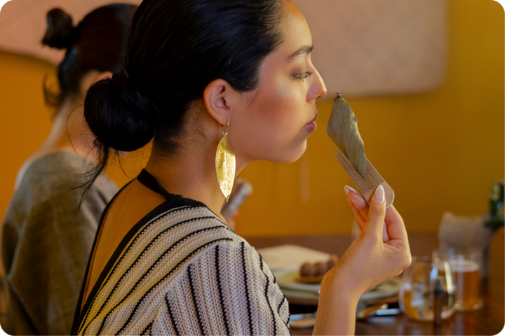 Un plan rapproché d'une personne tenant une feuille de laurier, assise à une table où sont posés des assiettes et des verres, évoquant une dégustation culinaire.
