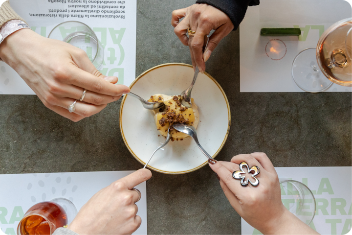 Plan rapproché sur les mains de quatre personnes partageant un dessert saupoudré de crumble, avec des boissons et des sets de table sur une table grise.