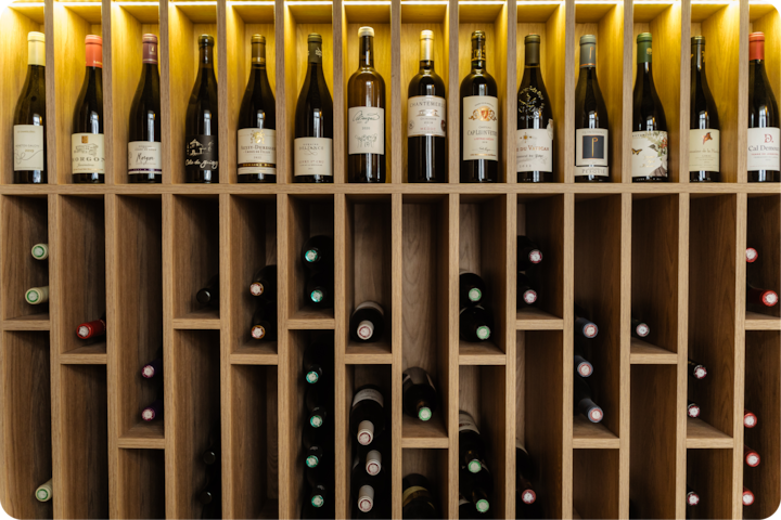 Des bouteilles de vin soigneusement disposées dans des casiers en bois, dont une rangée est éclairée par une lumière douce venant d'en haut et où les bouteilles sont debout avec leur étiquette bien visible.