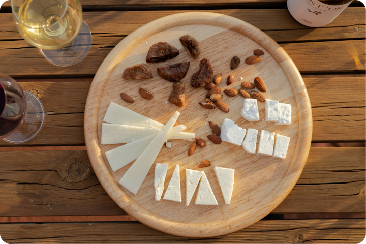 Un plateau rond en bois avec des fromages, des figues sèches et des amandes, avec à côté des verres de vin rouge et blanc, dans un cadre extérieur rustique.