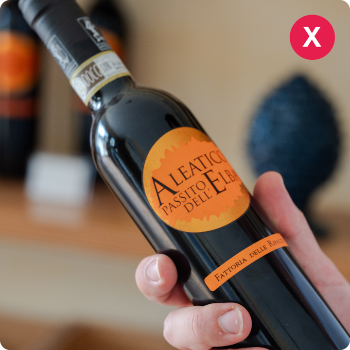 Plan rapproché d'une bouteille de vin étiquetée Aleatico Passito dell'Elba, dans la main d'une personne, avec une étagère et des objets flous en arrière-plan.