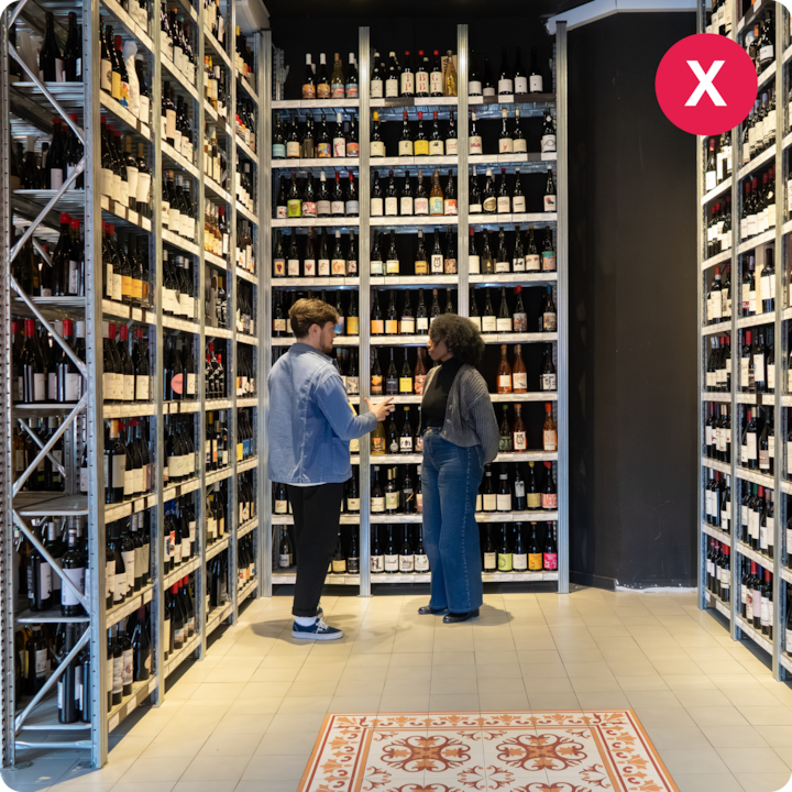 Deux personnes debout discutent dans une cave à vin, entourées d'étagères remplies de bouteilles.