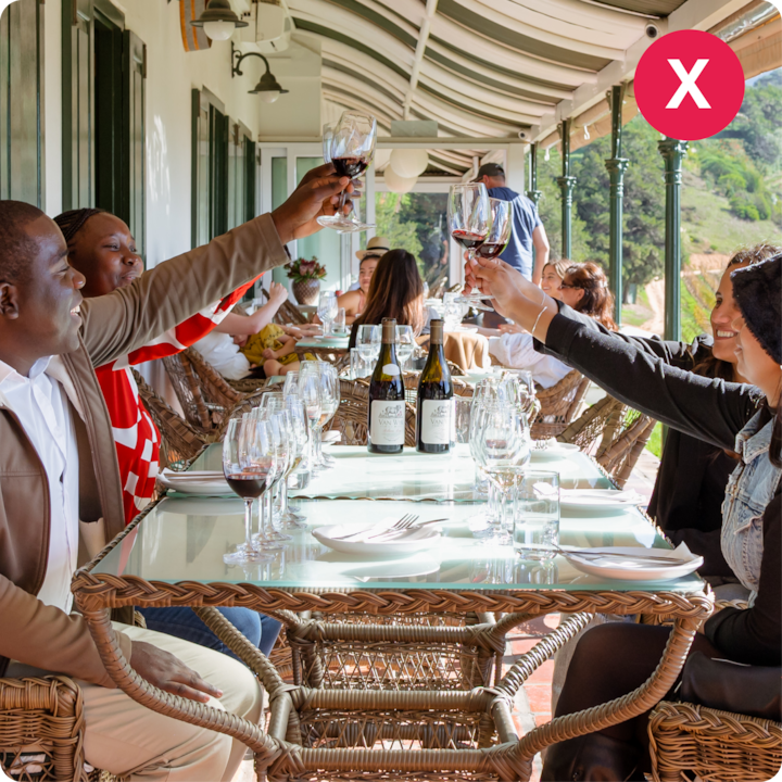 Une expérience de dégustation de vin sur une terrasse ensoleillée, avec des personnes levant leur verre de vin rouge, installées sur des chaises en rotin et autour d'une table en rotin où sont posés des bouteilles de vin et des verres.