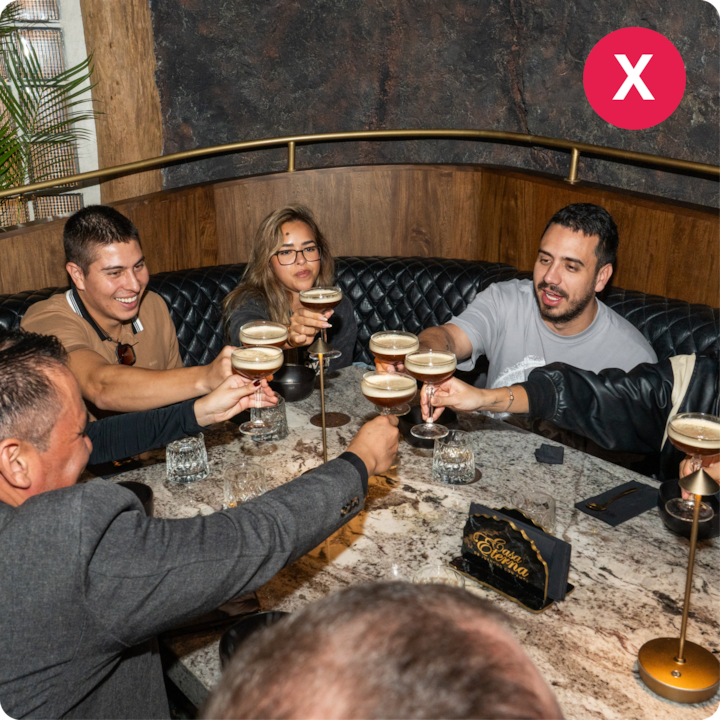 Des personnes assises autour d'une table en marbre portent un toast avec des coupes contenant un cocktail crémeux, dans un lieu confortable et faiblement éclairé, décoré de bois et de cuir.