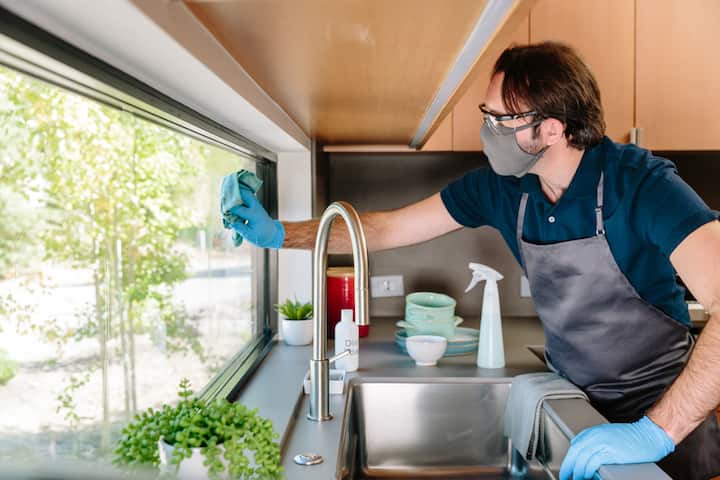 A person wearing gloves and an apron cleaning a kitchen window, with cleaning supplies and plants on the counter nearby, illustrating safety and hygiene practices for hosts.