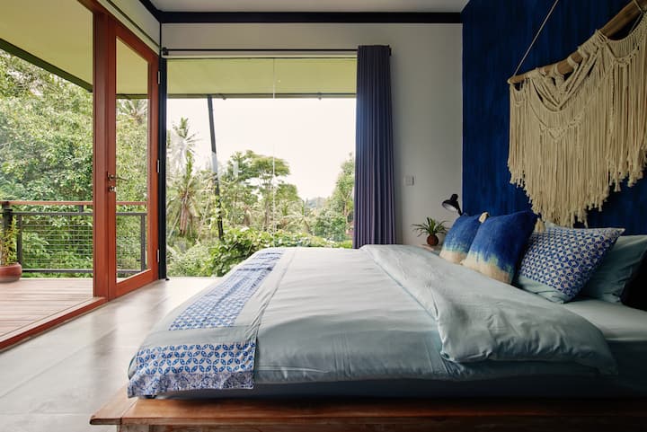Bright bedroom with a blue accent wall, large bed with patterned pillows and blanket, and a view of lush greenery through glass doors opening to a balcony.
