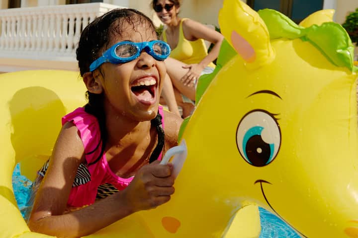 Child wearing a pink swimsuit playing on a yellow inflatable pool toy shaped like a dragon, with water splashing in a nearby pool and a sunny atmosphere.
