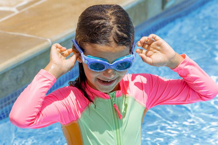 Child wearing colorful swim gear adjusts goggles near the edge of a pool, emphasizing pool safety for families with young children.