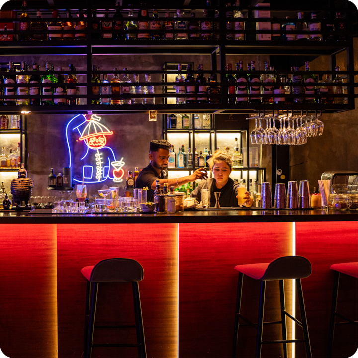 Un bar avec des panneaux au néon, des bouteilles soigneusement disposées sur des étagères et un comptoir éclairé en rouge. L'atmosphère évoque un lieu de vie nocturne tendance et décontracté.