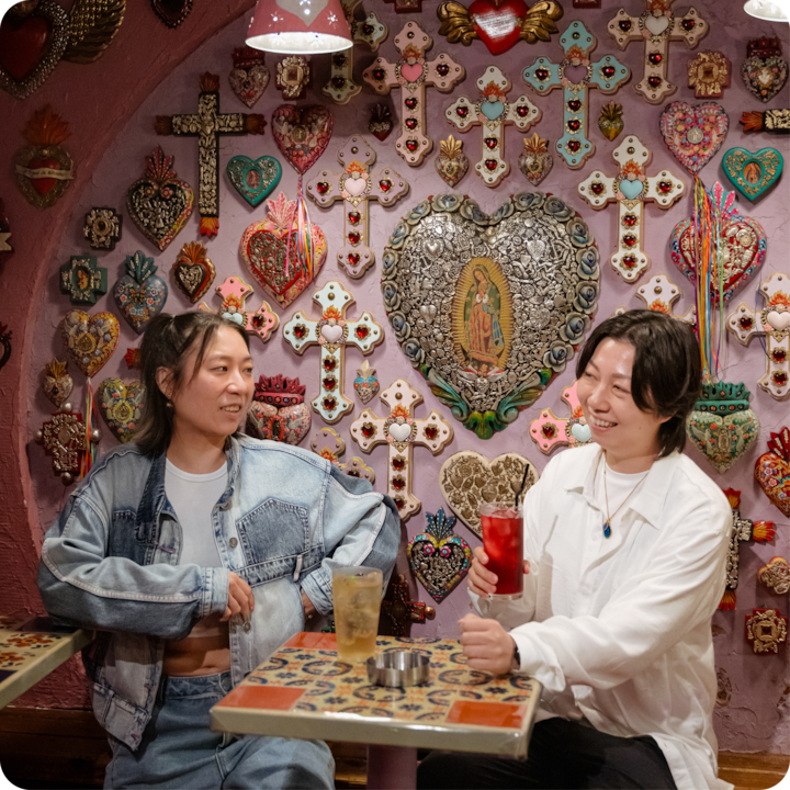 Un mur coloré orné de décorations religieuses, notamment des croix, des cœurs et une Vierge de Guadalupe au centre, avec deux personnes assises à une table carrelée en train de boire un verre.