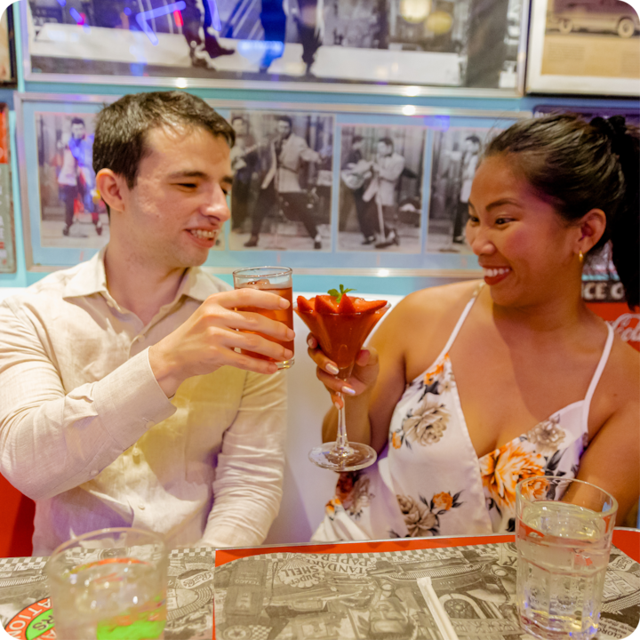 Ambiance de dîner décontractée où deux personnes trinquent avec des cocktails, entourées d'une décoration colorée comprenant des affiches et des photos vintage sur un mur bien éclairé.