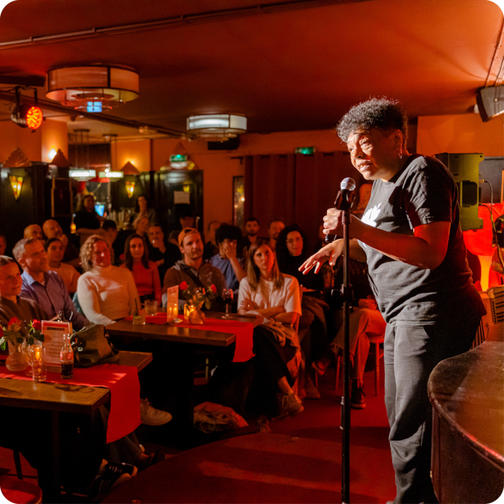 Un humoriste se produit sur scène dans un lieu faiblement éclairé par une lumière orange chaude, entouré d'un public attentif assis à des tables avec des boissons et des décorations.