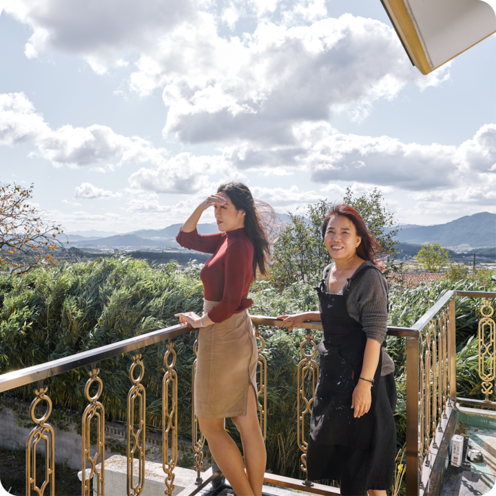 Deux personnes se tiennent sur un balcon avec des montagnes pittoresques, une végétation luxuriante et un ciel nuageux en arrière-plan, mettant en valeur l'éclairage d'un cadre extérieur.