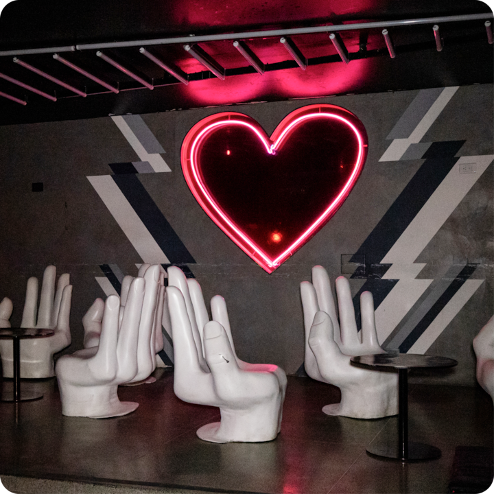 Un espace artistique avec des chaises blanches en forme de mains disposées près des tables, un néon cœur lumineux et des motifs géométriques sur les murs sous une lumière tamisée.