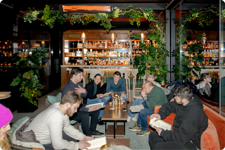 Un petit groupe de personnes dans un bar à l'éclairage chaleureux, décoré de miroirs et de plantes vertes, discutent ou examinent des documents autour d'une table.