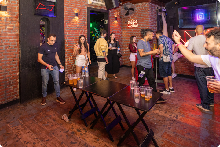 Une scène animée de fête en intérieur, avec des groupes de personnes discutant près d'une table dressée pour un jeu à boire, éclairée par des néons colorés sur des murs en briques.