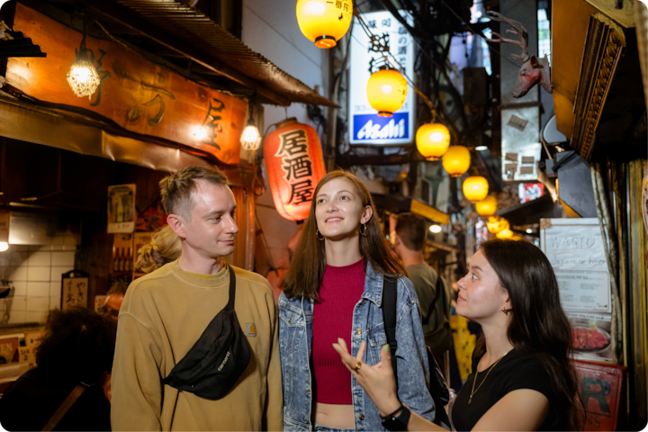 Une ruelle animée de nuit, éclairée par des lanternes suspendues. Des personnes sont rassemblées près de restaurants japonais traditionnels et d'enseignes colorées.