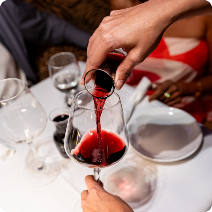 Un gros plan d'une main versant du vin rouge dans un verre sur une table dressée pour le dîner dans un cadre intérieur faiblement éclairé.