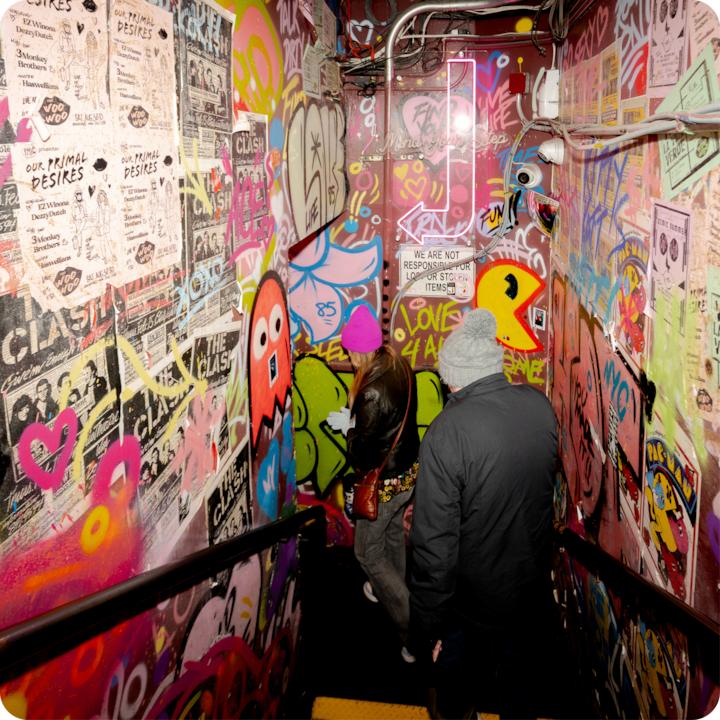 Un escalier faiblement éclairé recouvert de graffitis, d'affiches et d'œuvres de street art colorées, où descendent deux personnes, l'une portant un bonnet rose vif et l'autre un couvre-chef gris.