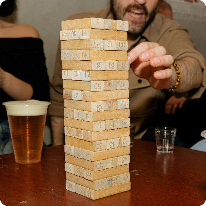 Une personne attrape avec précaution un bloc d'un jeu empilable en bois sur une table dans un espace intérieur faiblement éclairé, avec des boissons visibles à proximité.