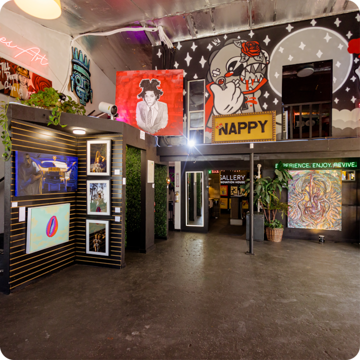 L'intérieur d'un espace créatif présentant une diversité artistique avec des peintures murales, des œuvres d'art encadrées, des enseignes au néon et une végétation luxuriante. Un mélange d'éclairages et de décorations colorés crée une ambiance ludique.