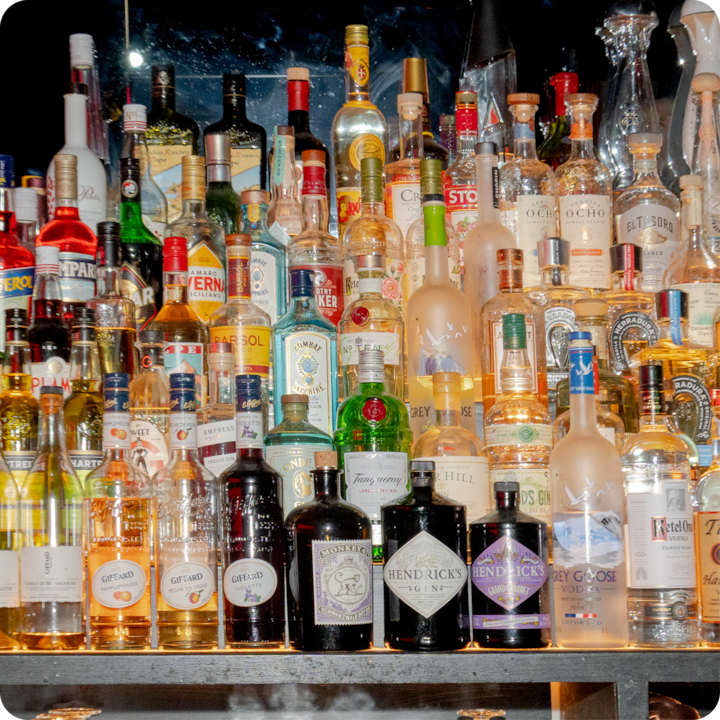 Des étagères remplies de diverses bouteilles d'alcool, illuminées par des néons et un éclairage d'ambiance, soulignant les couleurs riches et les motifs complexes des bouteilles.