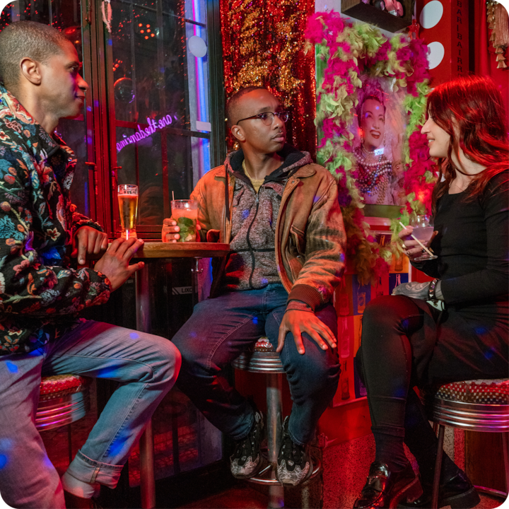 Un cadre accueillant dans un bar aux murs colorés, éclairé par des néons et orné de décorations éclatantes. Des boissons sont disposées sur la table et les clients échangent dans une ambiance conviviale et chaleureuse.