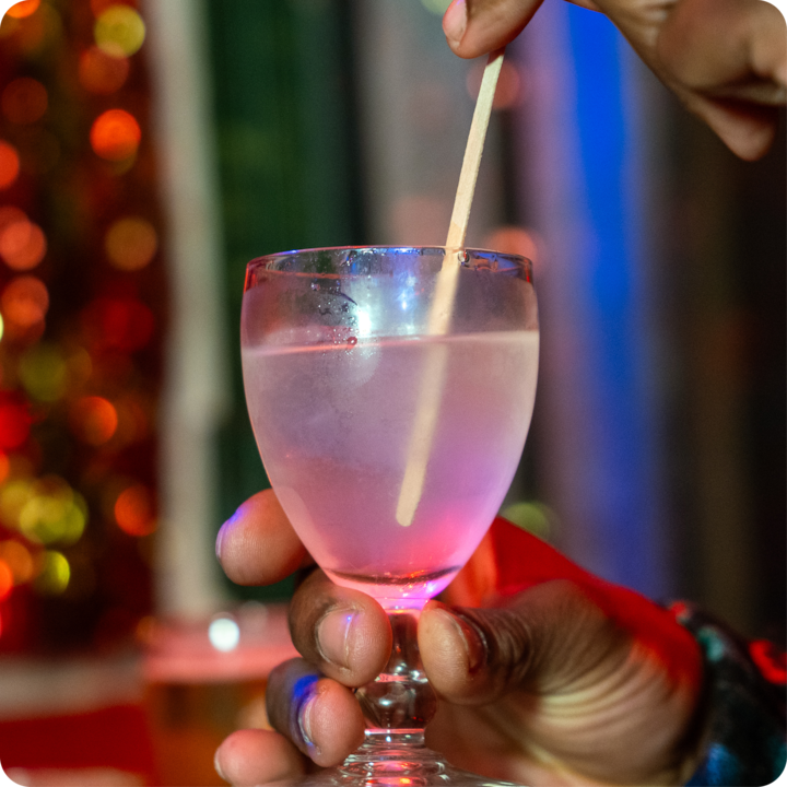 Gros plan sur une personne remuant une boisson rose dans un verre, sur fond d'éclairage d'ambiance coloré présentant des nuances floues de rouge et de bleu.