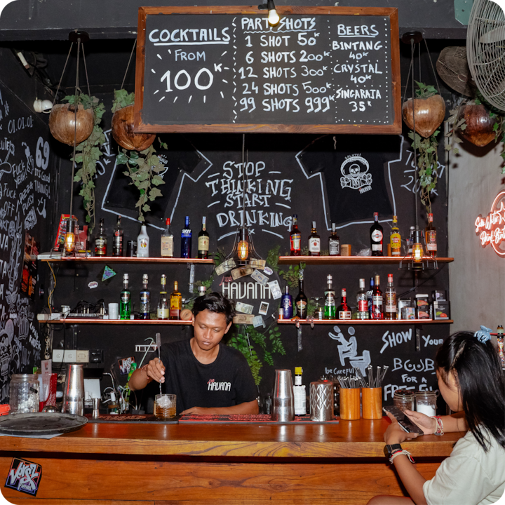 Un bar chaleureux de nuit, avec un barman préparant une boisson sous un tableau noir affichant les prix des cocktails, des shots et des bières. Les étagères du bar sont garnies de bouteilles d'alcool et entourées de plantes suspendues, d'enseignes au néon et de dessins à la craie.