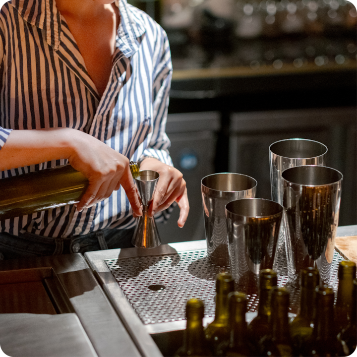 Dans la pénombre du soir, une personne prépare des boissons dans un bar à l'aide d'un doseur. Autour d'elle se trouvent des shakers à cocktails et des bouteilles.