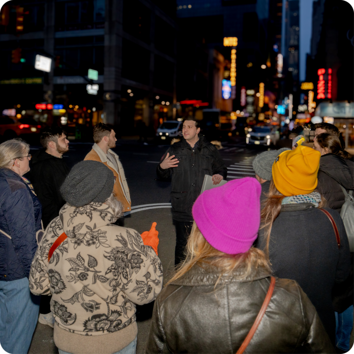 Scène nocturne de rue dans une ville avec un petit groupe de personnes interagissant sous un éclairage artificiel. Les environs sont composés d'enseignes et de bâtiments éclairés, soulignant l'environnement urbain nocturne.