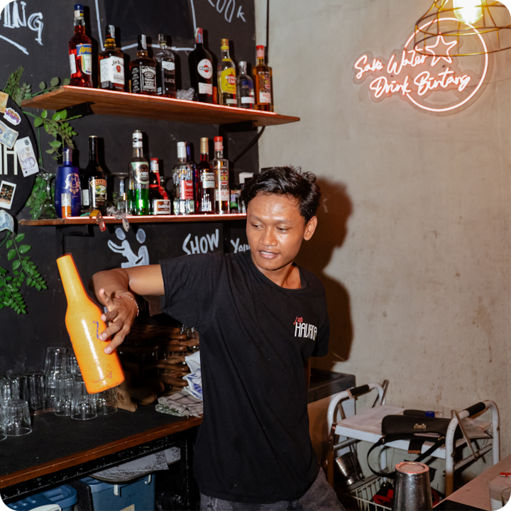 Un bar animé avec un barman en action, des verres, des bouteilles d'alcool exposées et une enseigne au néon qui ajoute de l'ambiance à l'environnement faiblement éclairé.