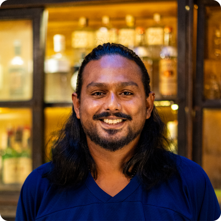 Un portrait d'une personne vêtue d'un t-shirt bleu, pris dans une ambiance tamisée, avec en arrière-plan des étagères remplies de bouteilles et d'accessoires de bar, éclairées par une lumière douce.
