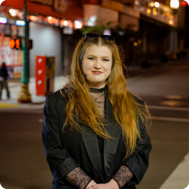 Une personne portant une tenue noire avec des détails en dentelle se tient dans une rue peu éclairée avec des vitrines illuminées et un environnement urbain en arrière-plan.