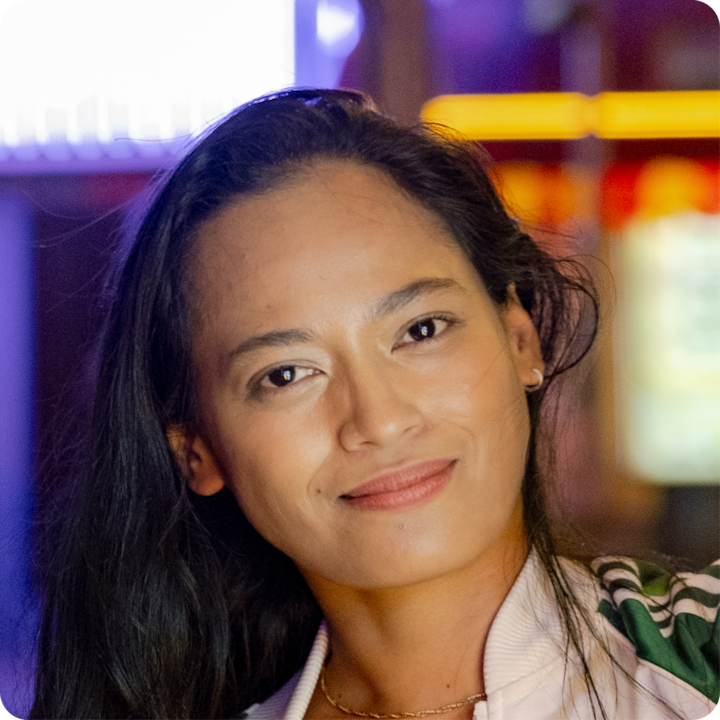 Une personne aux longs cheveux noirs, portant une veste blanche avec des touches de vert, est photographiée dans un environnement faiblement éclairé, avec à l'arrière-plan des lumières éclatantes et floues.