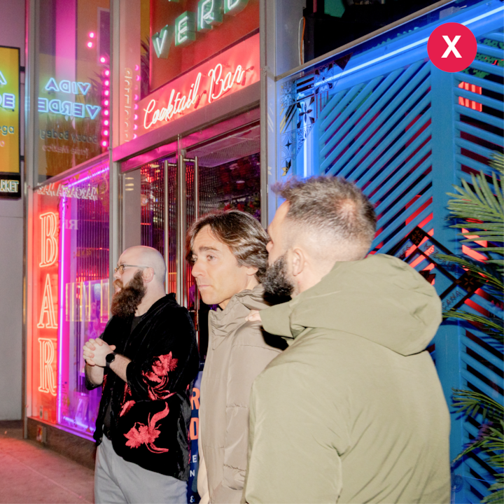 Trois personnes se tiennent près de l'entrée d'un bar à cocktails illuminée par des néons la nuit, illustrant un mélange de sources lumineuses.