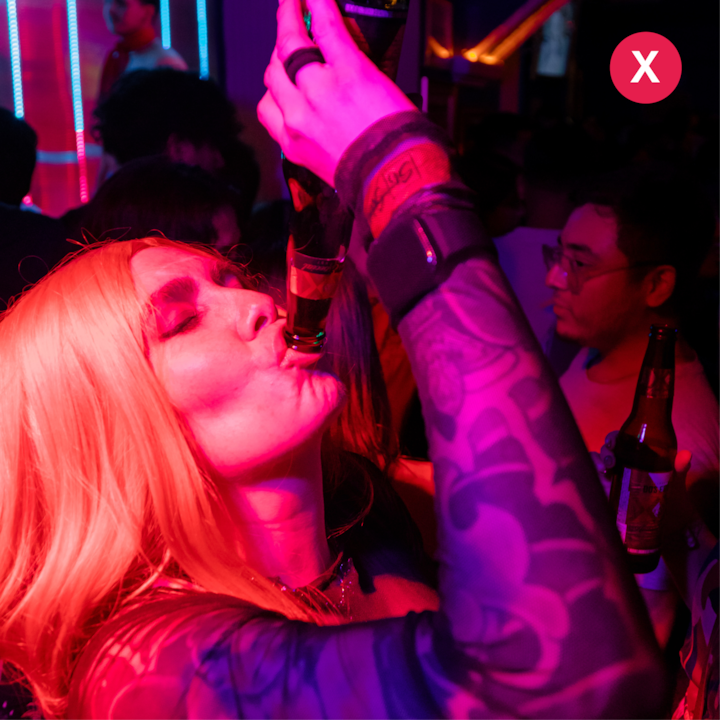 Une scène de boîte de nuit faiblement éclairée, avec des lumières rouges et violettes, montrant une personne avec un tatouage sur tout le bras buvant à la bouteille, entourée d'autres personnes tenant des boissons.