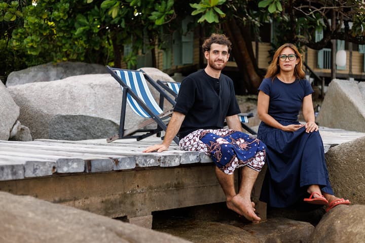 Two people sitting on a wooden dock surrounded by large rocks and lush greenery, with striped deck chairs in the background.