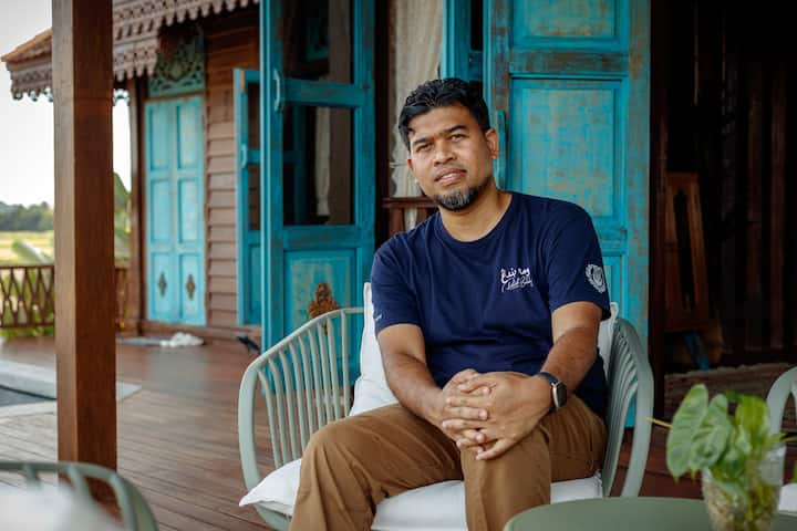 A person seated on a patio in Langkawi, surrounded by vibrant blue wooden doors and traditional Malaysian architecture.