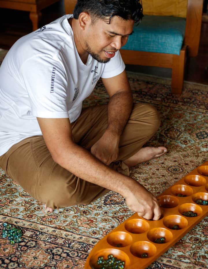 A person sitting cross-legged on a patterned rug, playing a traditional wooden mancala game with green glass beads.